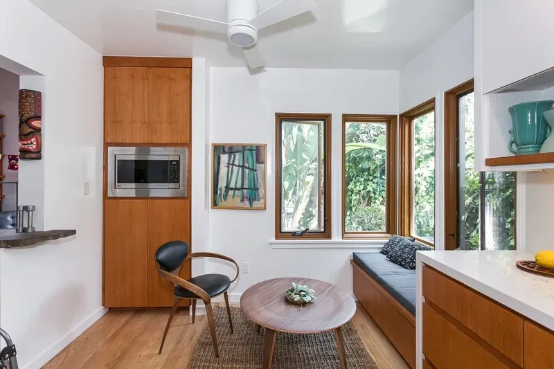 Cozy kitchen nook with wood cabinets, round table, and cushioned window seat overlooking greenery.