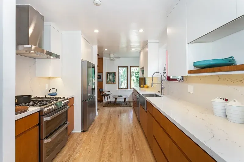 Modern kitchen with white countertops and wooden cabinets, stainless steel appliances, and a view into living area.