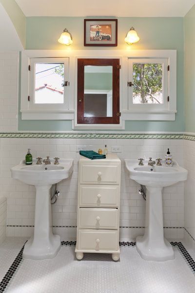 Bathroom with two pedestal sinks, center cabinet, and framed windows. Green and white tile, white walls.