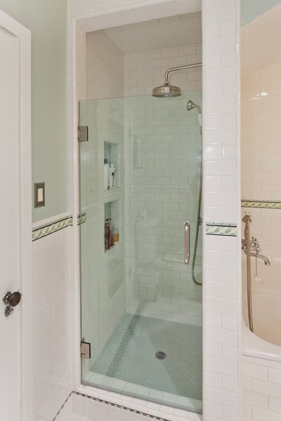 A clear glass shower with a recessed shelf, next to a wall with a light switch and tiled trim.