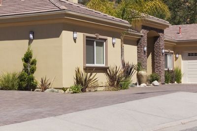 Tan stucco house with plants, brick accents, and a paved driveway on a sunny day.