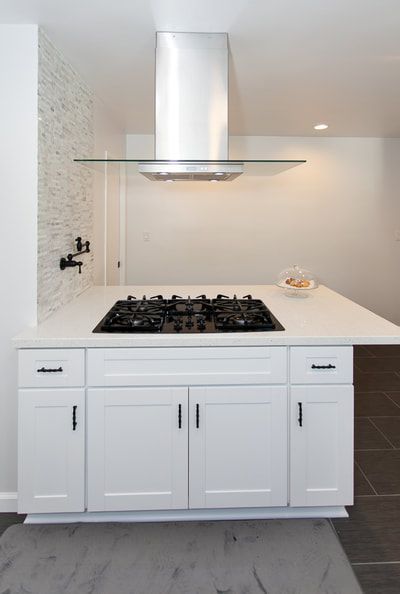 White kitchen island with a cooktop, range hood, and cabinets. Black accents, light gray floor.