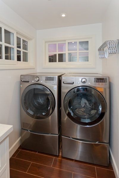 Laundry room with stainless steel washer and dryer, wood-look tile floor, white walls, windows.