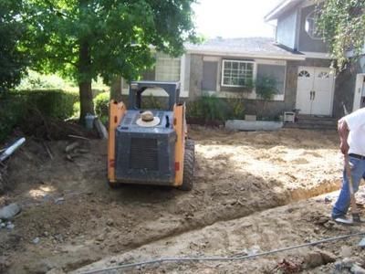 A small orange skid steer and a man digging in a dirt yard near a house.