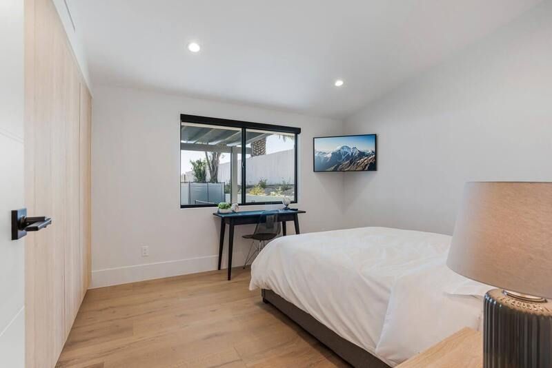 Bedroom with bed, desk by window, TV on wall, and light-colored wood floor.