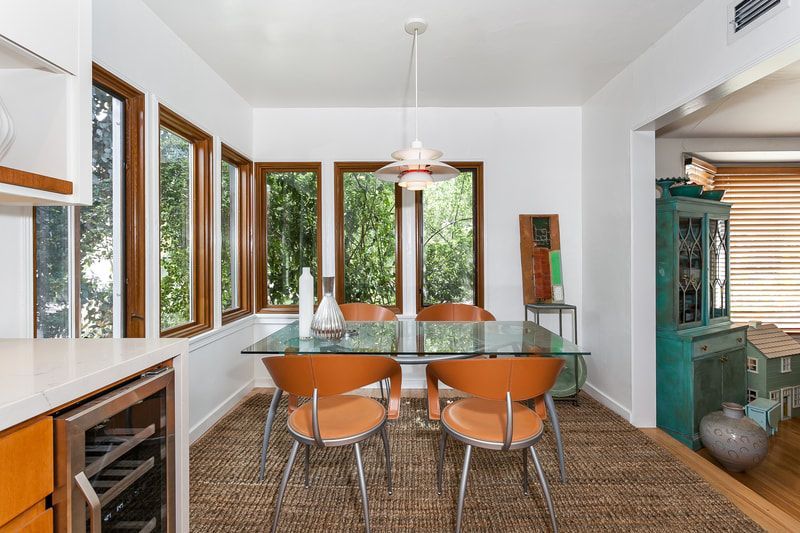 Dining room with glass table, orange chairs, and windows overlooking greenery.
