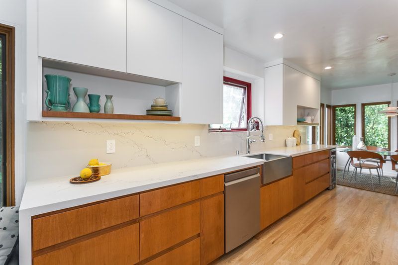 Modern kitchen with light wood cabinets, white countertops, stainless steel sink, and a view into a dining area.