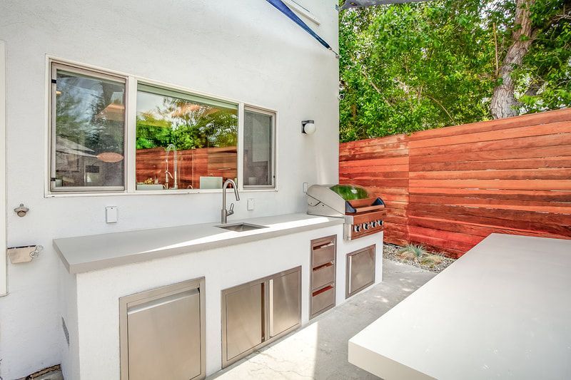 Outdoor kitchen with a sink, grill, and cabinets against a white wall, next to a wooden fence.