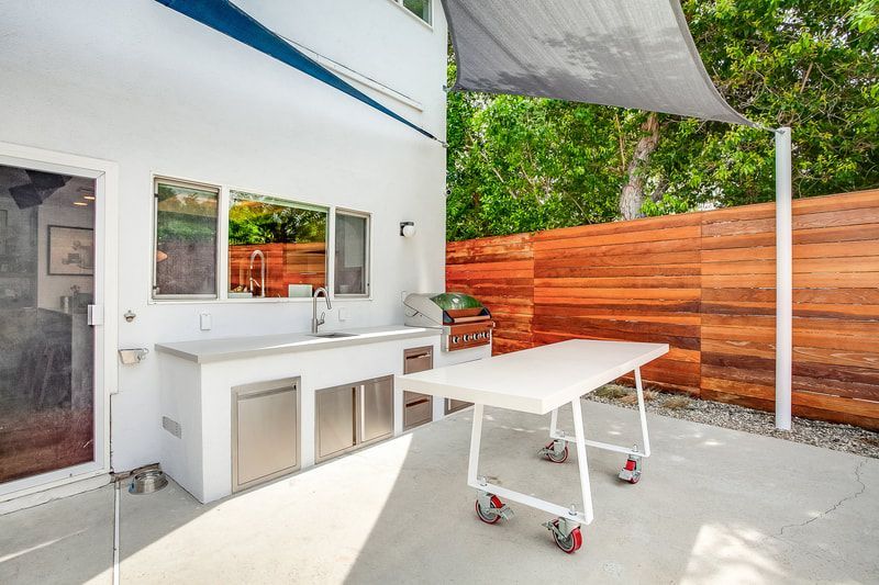 Outdoor kitchen with grill, sink, and table, next to a wooden fence and green trees.