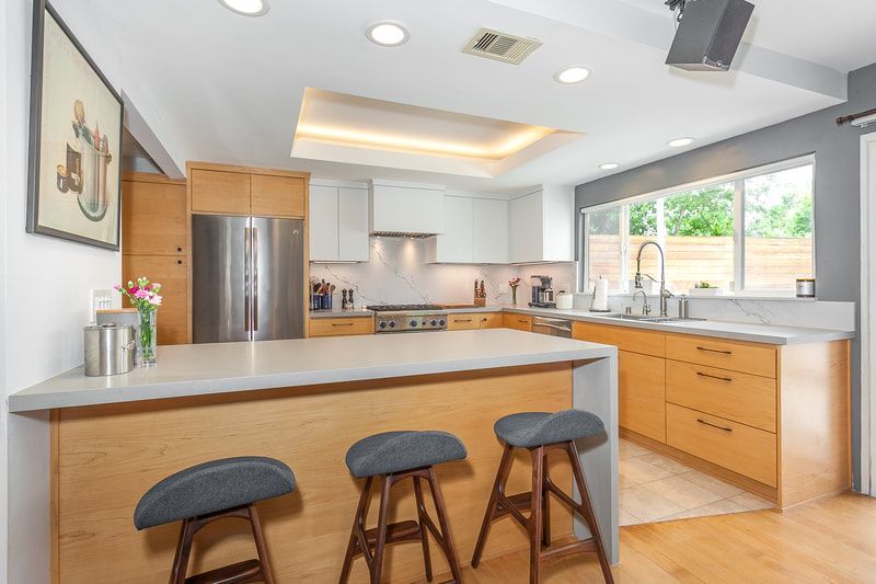 Modern kitchen with light wood cabinets, gray countertops, and three gray stools at the island.