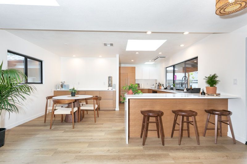 Bright, modern kitchen with wood and white accents; bar stools at counter, dining table, and plants.