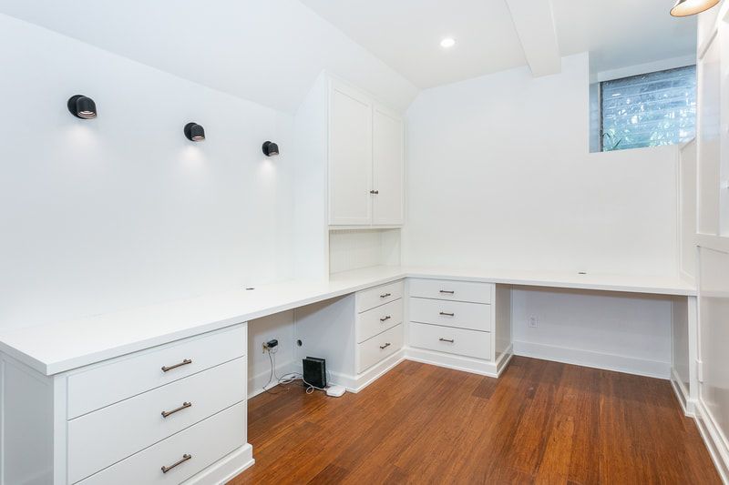 White office with built-in desks, drawers, and cabinets, hardwood floor, and three black sconces on the wall.