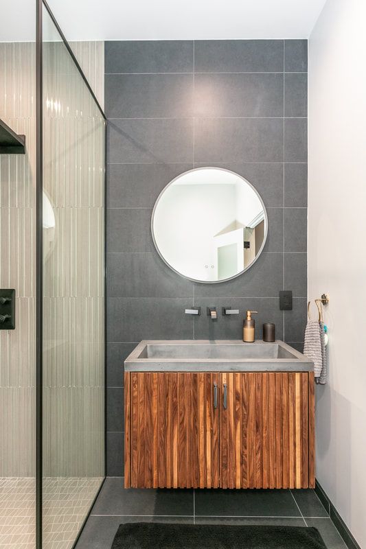 Modern bathroom: wooden vanity with concrete sink, round mirror, gray tiled wall, glass shower.