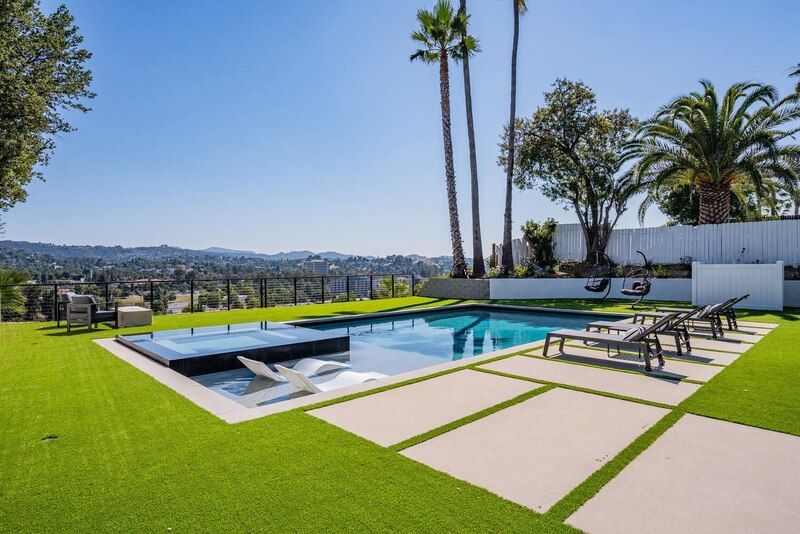 Pool with lounge chairs, artificial turf, and city view under a blue sky.