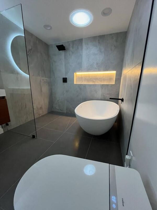 Modern bathroom with a white tub, gray tile, and a round mirror with ambient lighting.