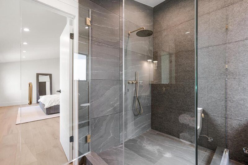 Modern bathroom with a glass-walled shower, gray tile, and an open doorway to a bedroom.