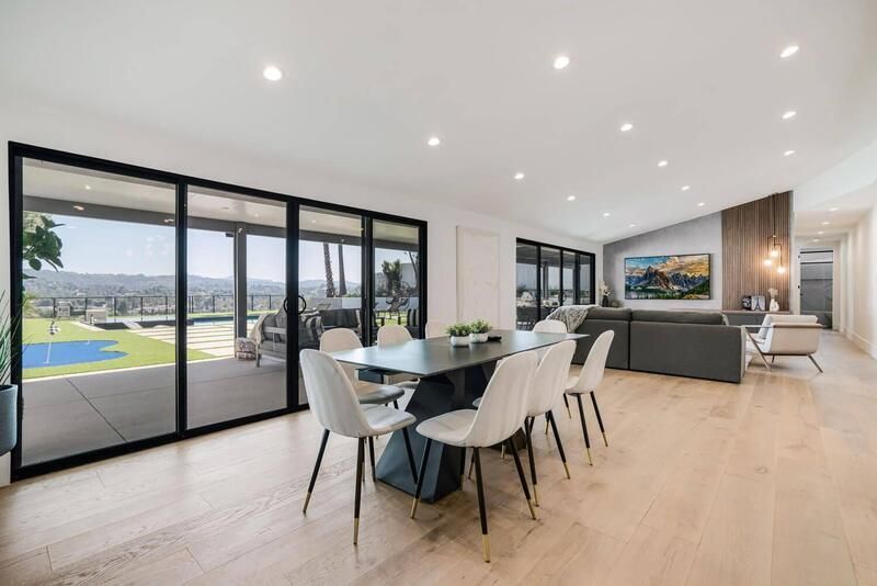 Modern dining and living space with large windows, overlooking a backyard, featuring a table, chairs, and a sofa.