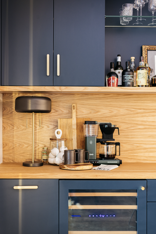 A modern home bar with navy cabinets, wood accents, and coffee station. Wine cooler and liquor bottles on display.