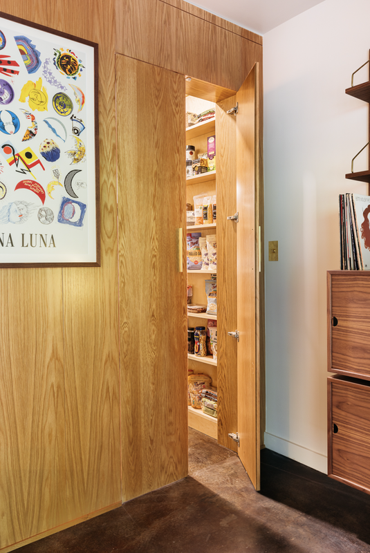 Wooden pantry door open, revealing shelves of food items. Oak paneling. Artwork on wall.