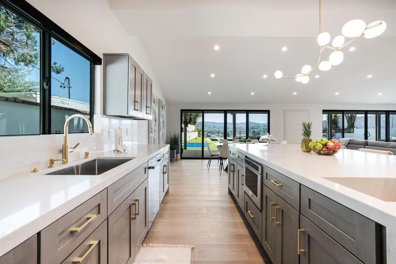 Modern kitchen with gray cabinets, white countertops, gold accents, and large windows overlooking a pool.