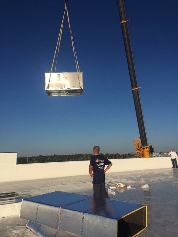 A crane lifting an HVAC unit onto a white rooftop, with a worker looking on
