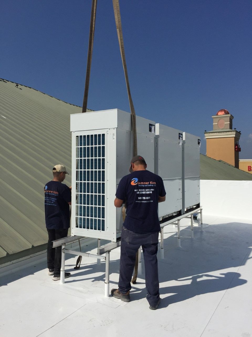 Two workers installing HVAC unit on a white rooftop, lifted by a crane