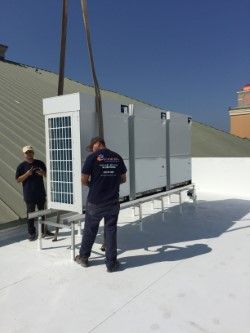 Two men on a white rooftop with a large HVAC unit being lifted by a crane