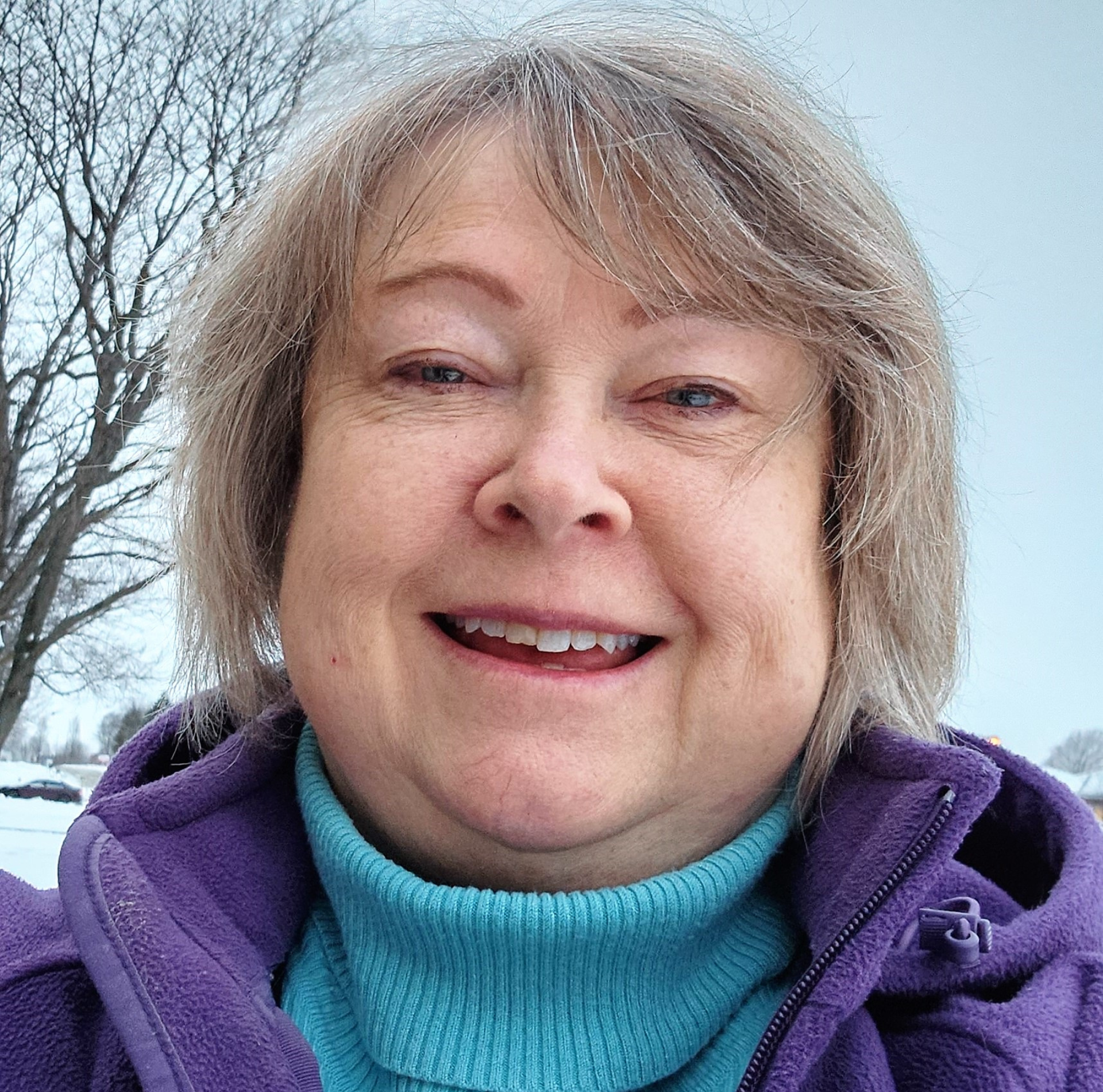 A woman wearing a purple jacket and a blue turtleneck is smiling.