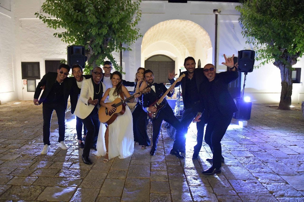 Gruppo musicale che si esibisce all'aperto di notte, di fronte a un edificio illuminato, con chitarre e sassofoni in un cortile di pietra.