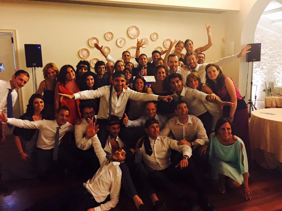 Un folto gruppo di persone in posa al chiuso, in abiti formali, sorridenti e con le mani alzate durante una celebrazione.