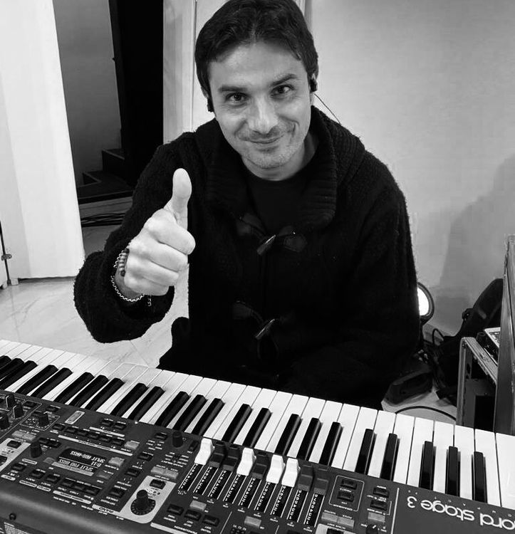 Un musicista sorridente fa il segno del pollice in su da dietro una tastiera, in una foto in bianco e nero.