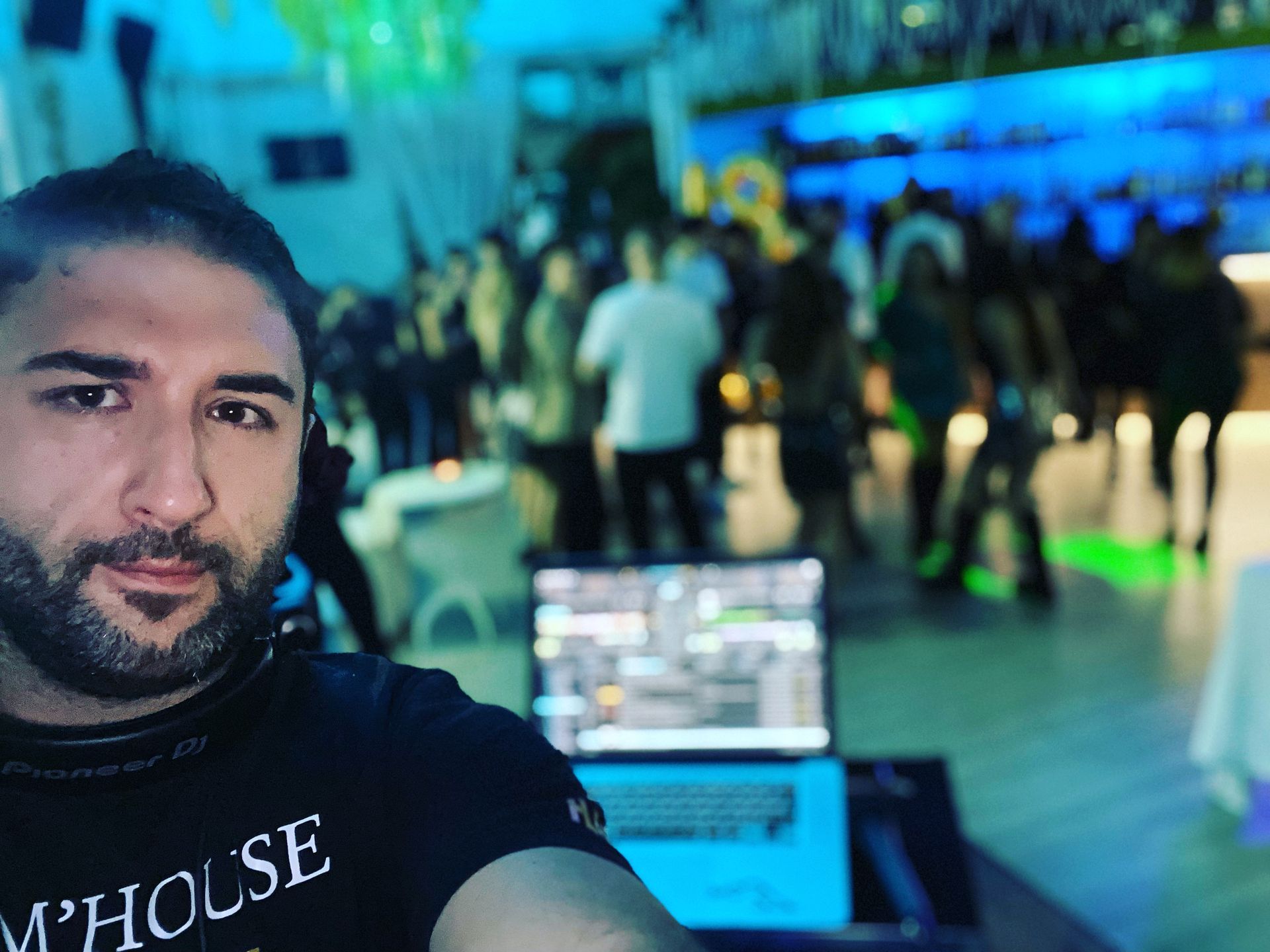 Un uomo si scatta un selfie in uno spazio eventi affollato e illuminato di blu, con un computer portatile in primo piano.