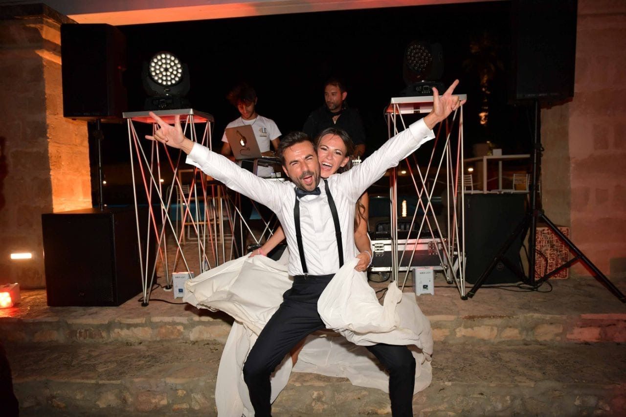 Uomo con le braccia alzate davanti alla consolle del DJ durante una festa notturna, con indosso una camicia bianca e le bretelle.