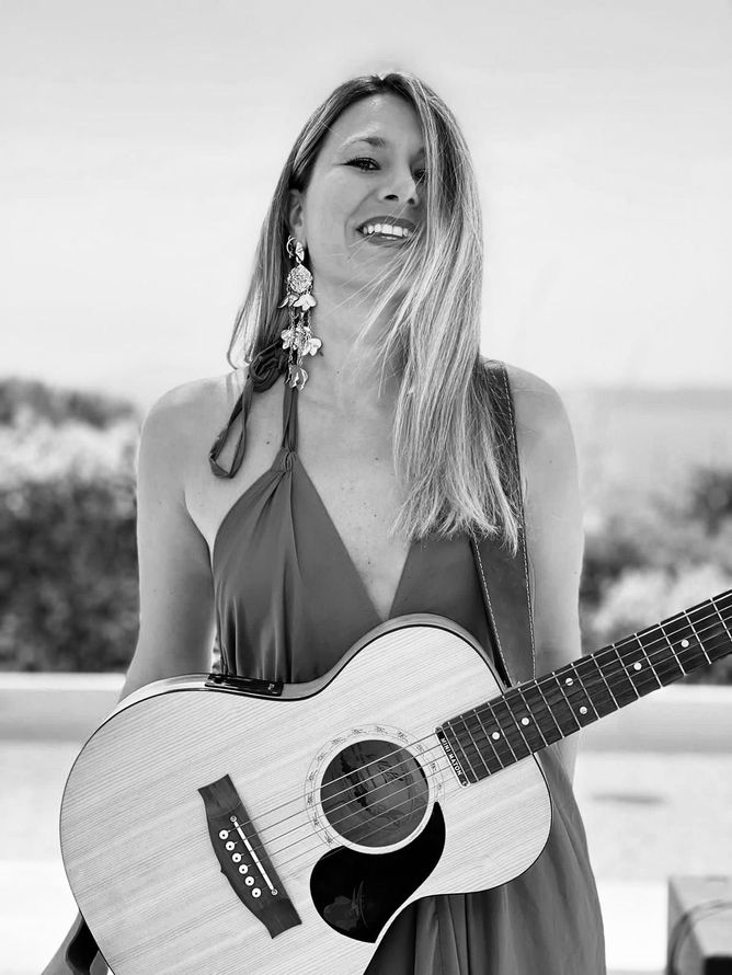 Donna sorridente all'aperto con una chitarra acustica, indossa un top scuro senza spalline.