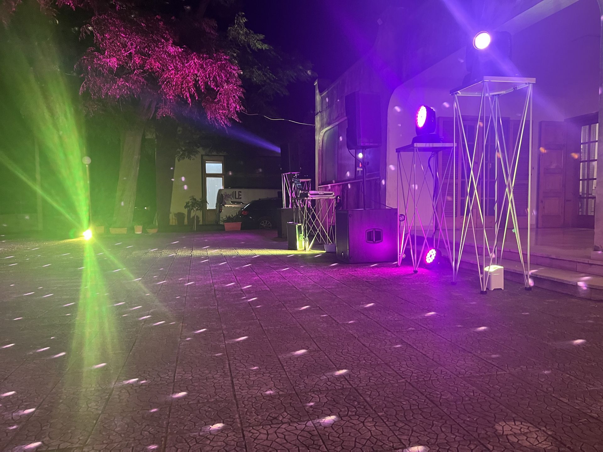 Luci laser viola e verdi su un patio di notte, con un edificio illuminato e alberi sullo sfondo.