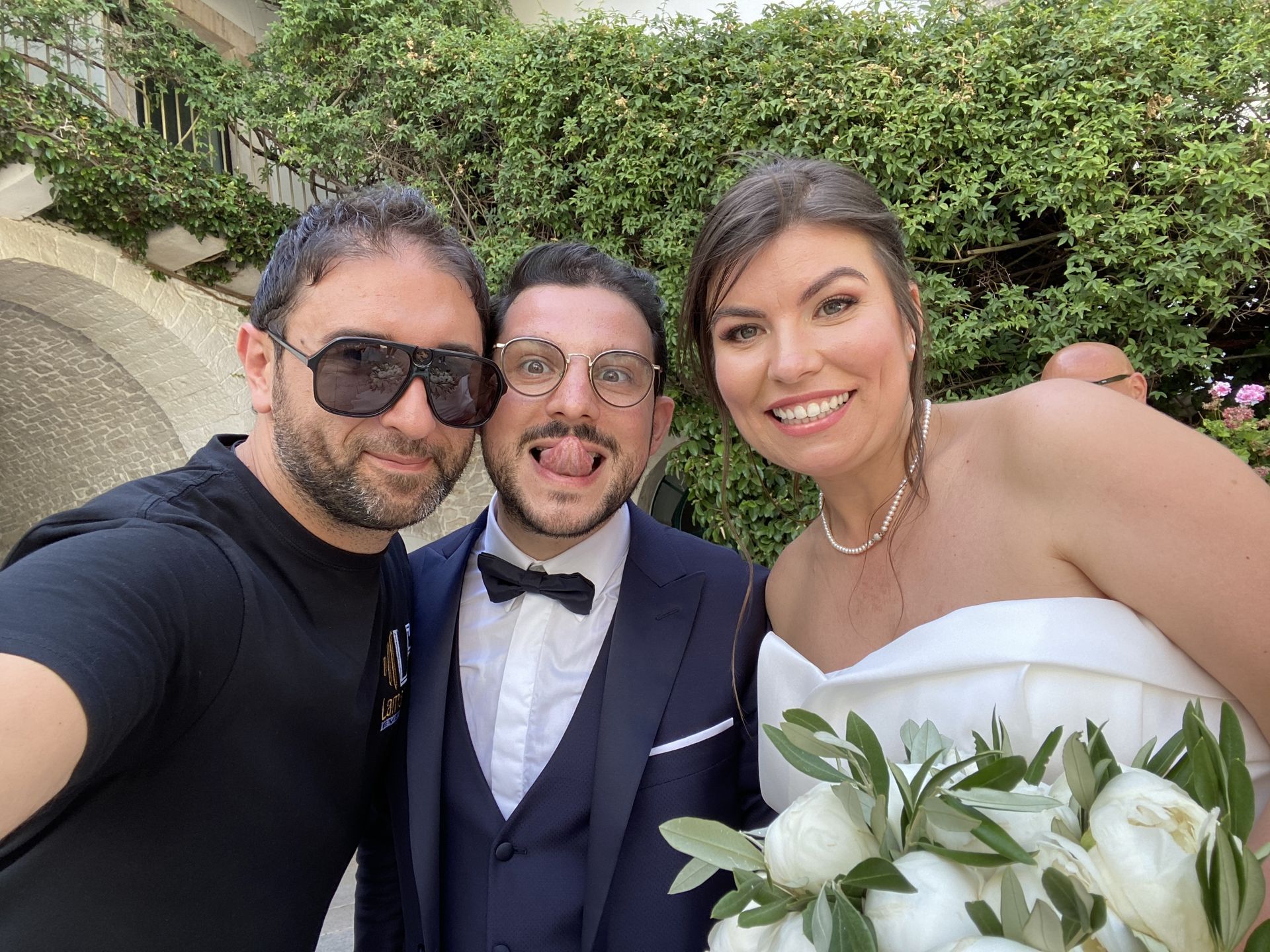 Tre persone sorridenti in posa all'aperto, una delle quali tiene in mano un bouquet bianco a un matrimonio.