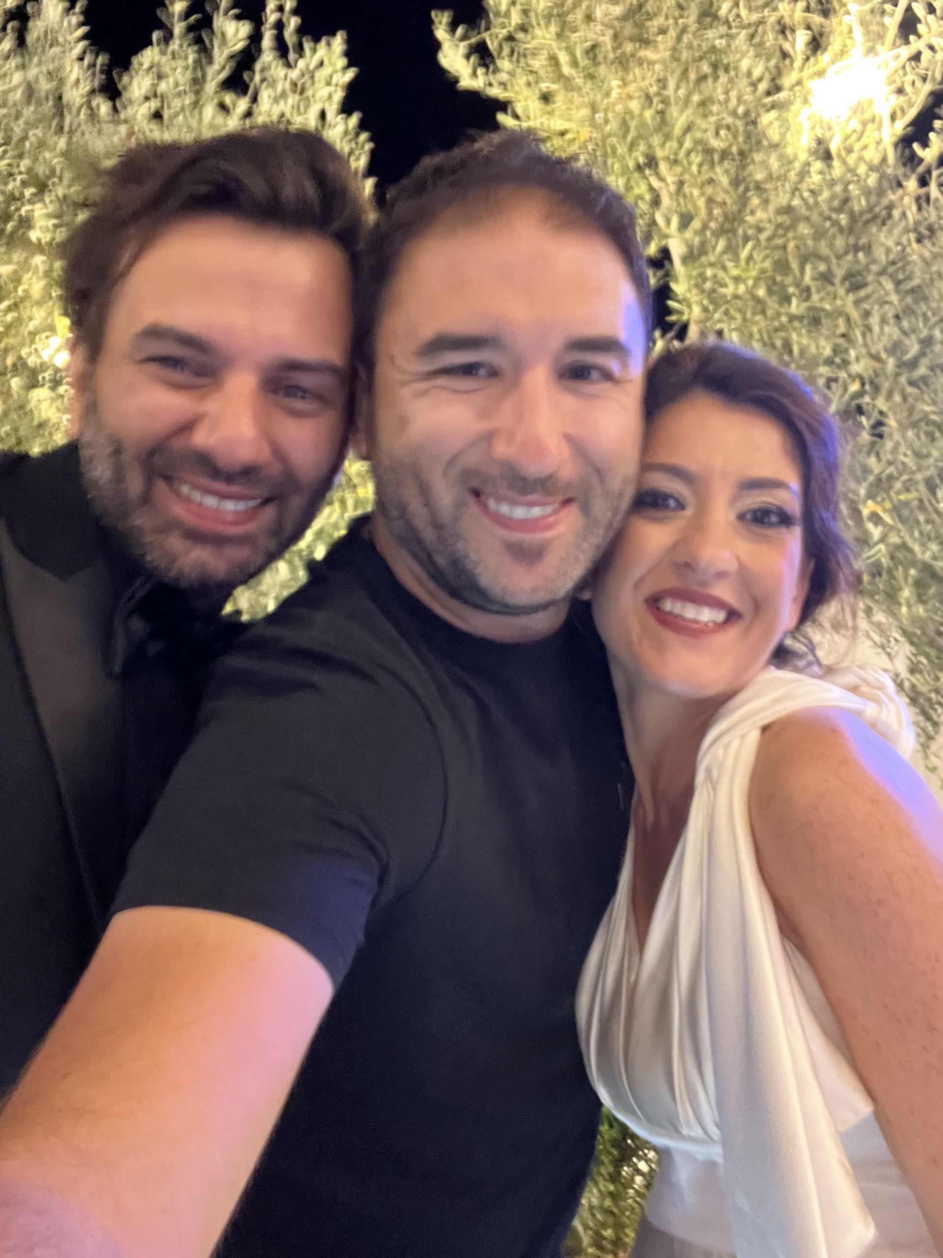 Tre amici sorridono vicini all'aperto di notte, con gli alberi che brillano sullo sfondo.