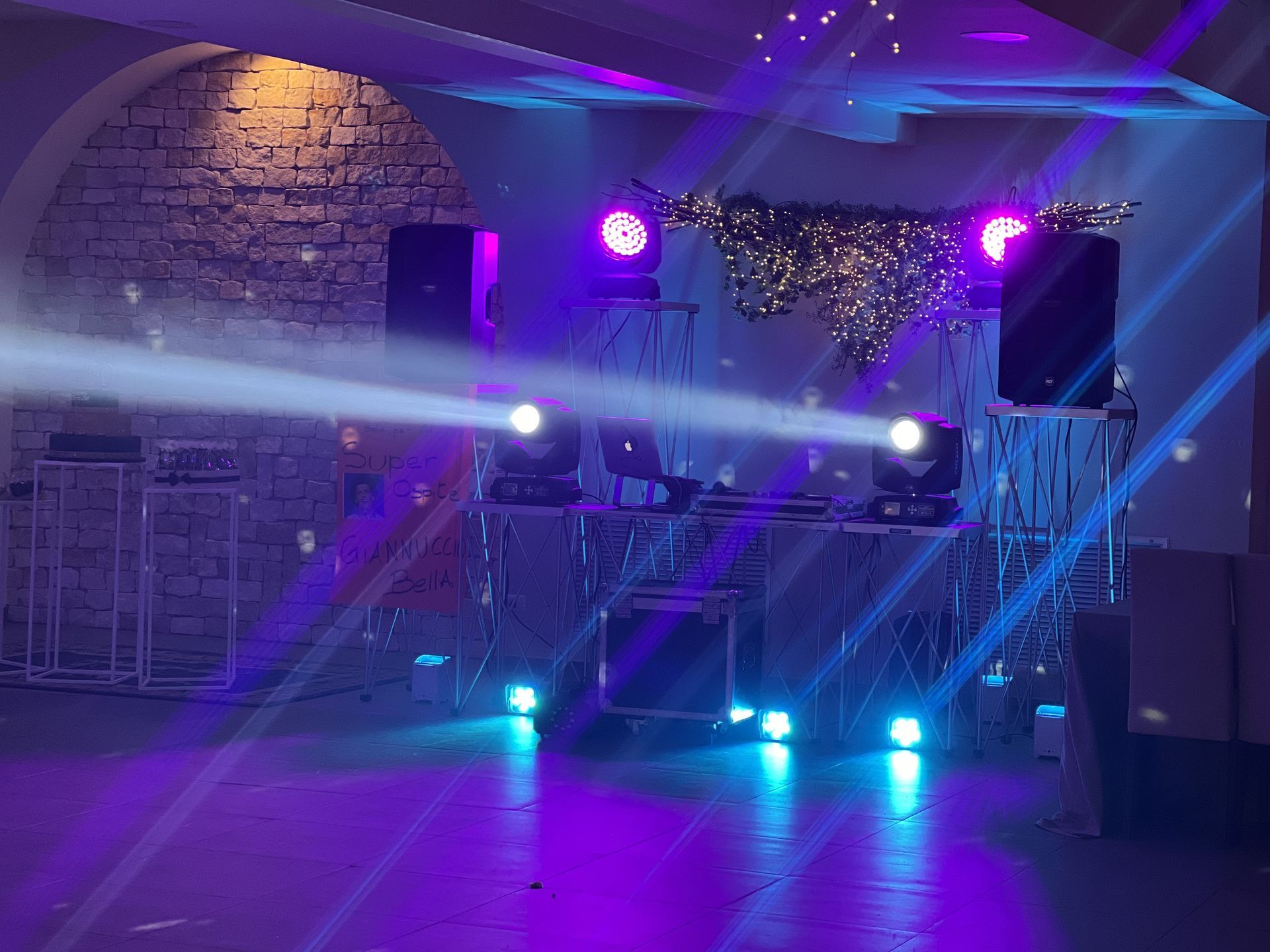 Palco illuminato di viola con fumo, riflettori brillanti e una postazione da DJ in una stanza con pareti in pietra.