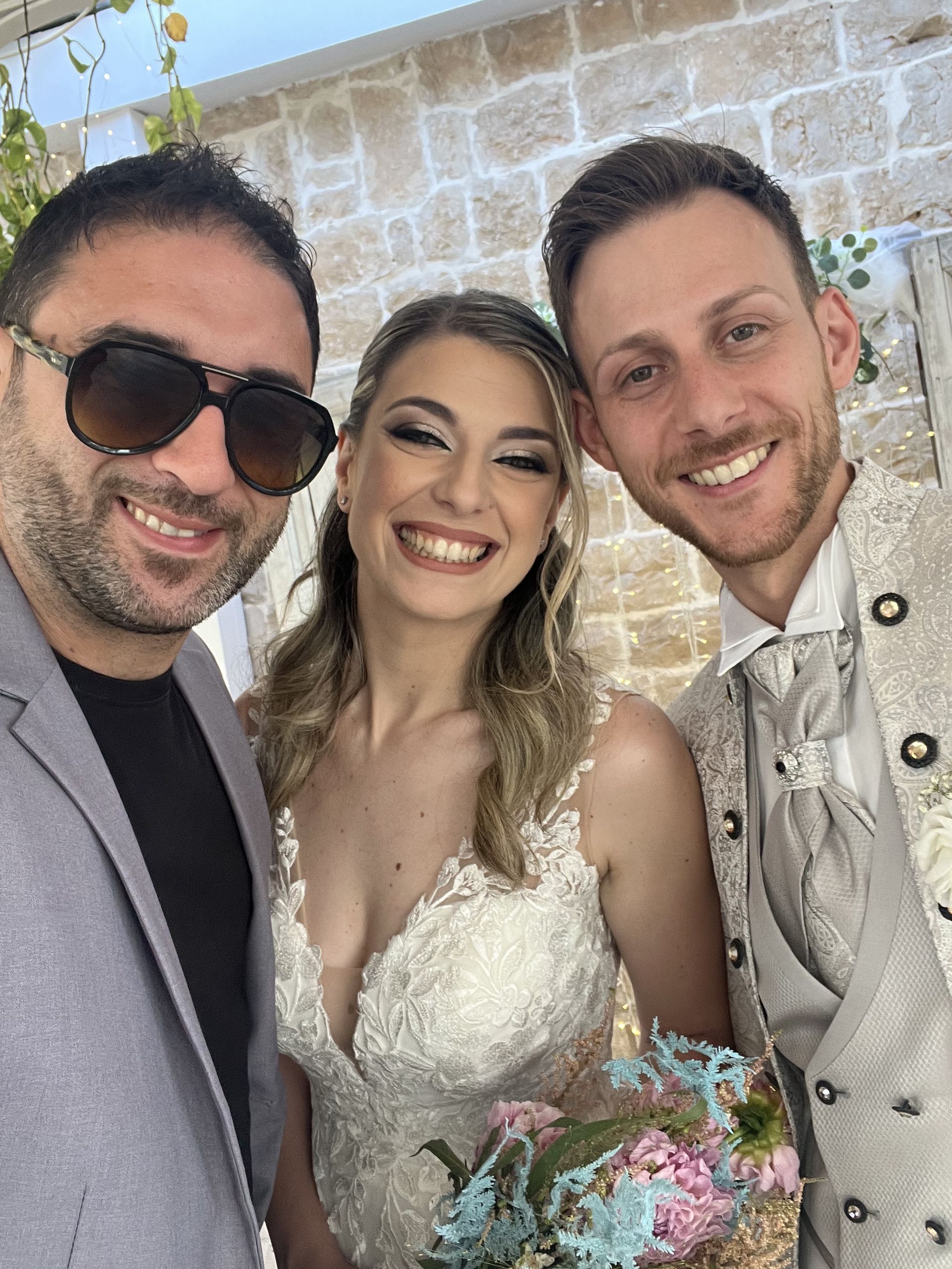 Tre persone sorridenti a un matrimonio, con la sposa in abito bianco che tiene in mano un bouquet dai colori pastello.