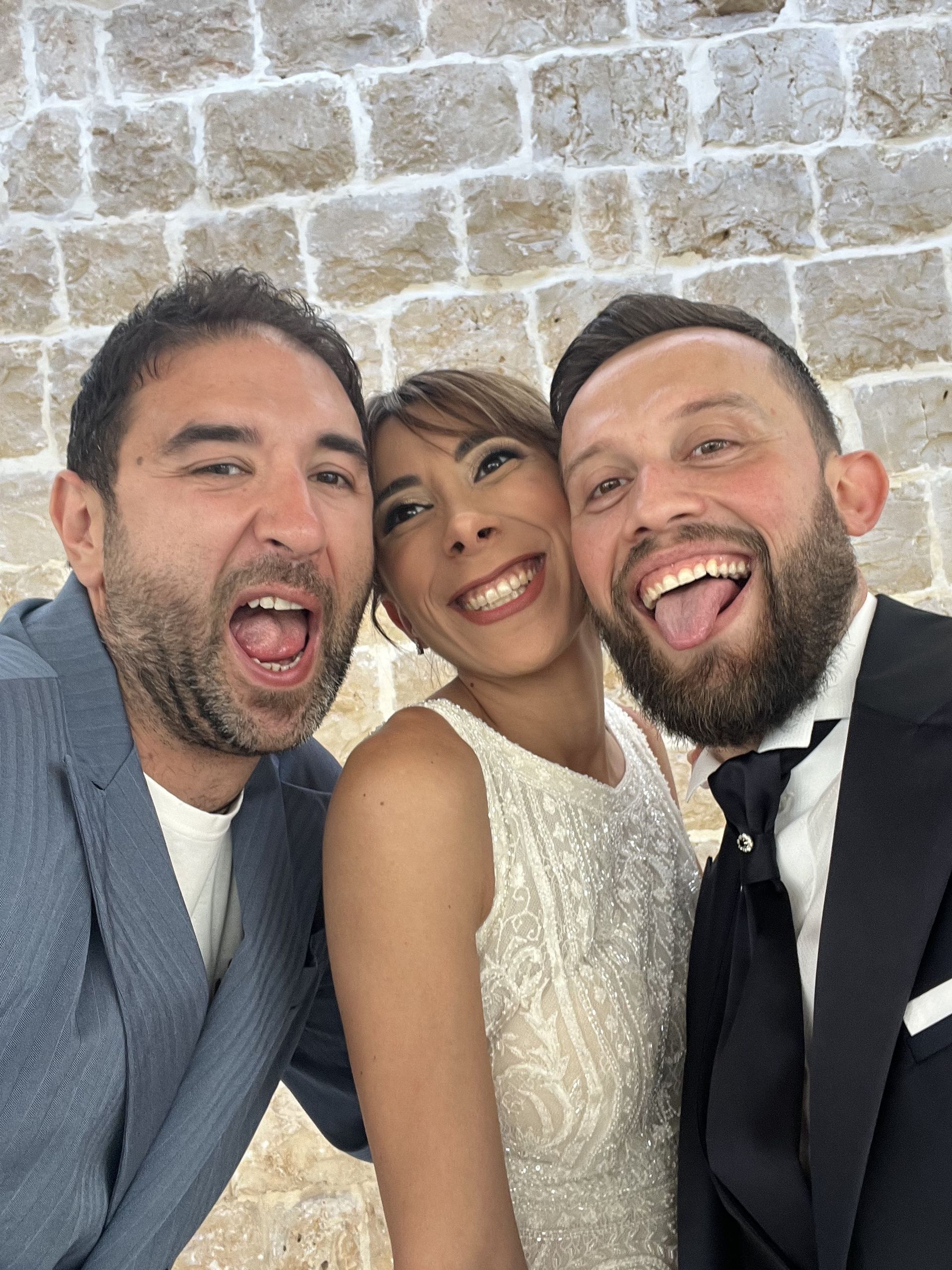 Tre persone sorridenti e in posa vicine davanti a un muro di pietra.