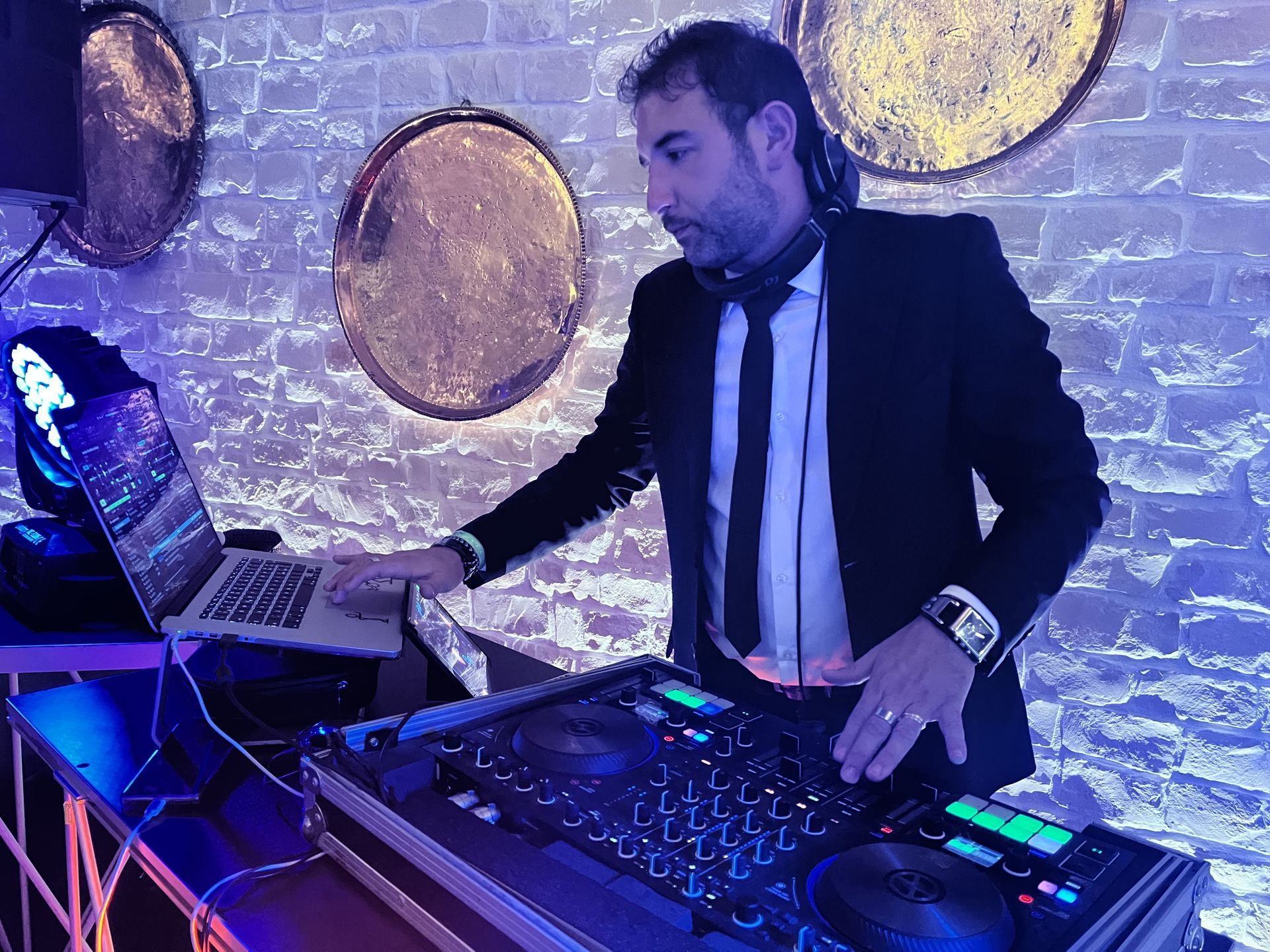 DJ che si esibisce con un laptop e un mixer in un locale illuminato di viola, con decorazioni murali circolari alle sue spalle.