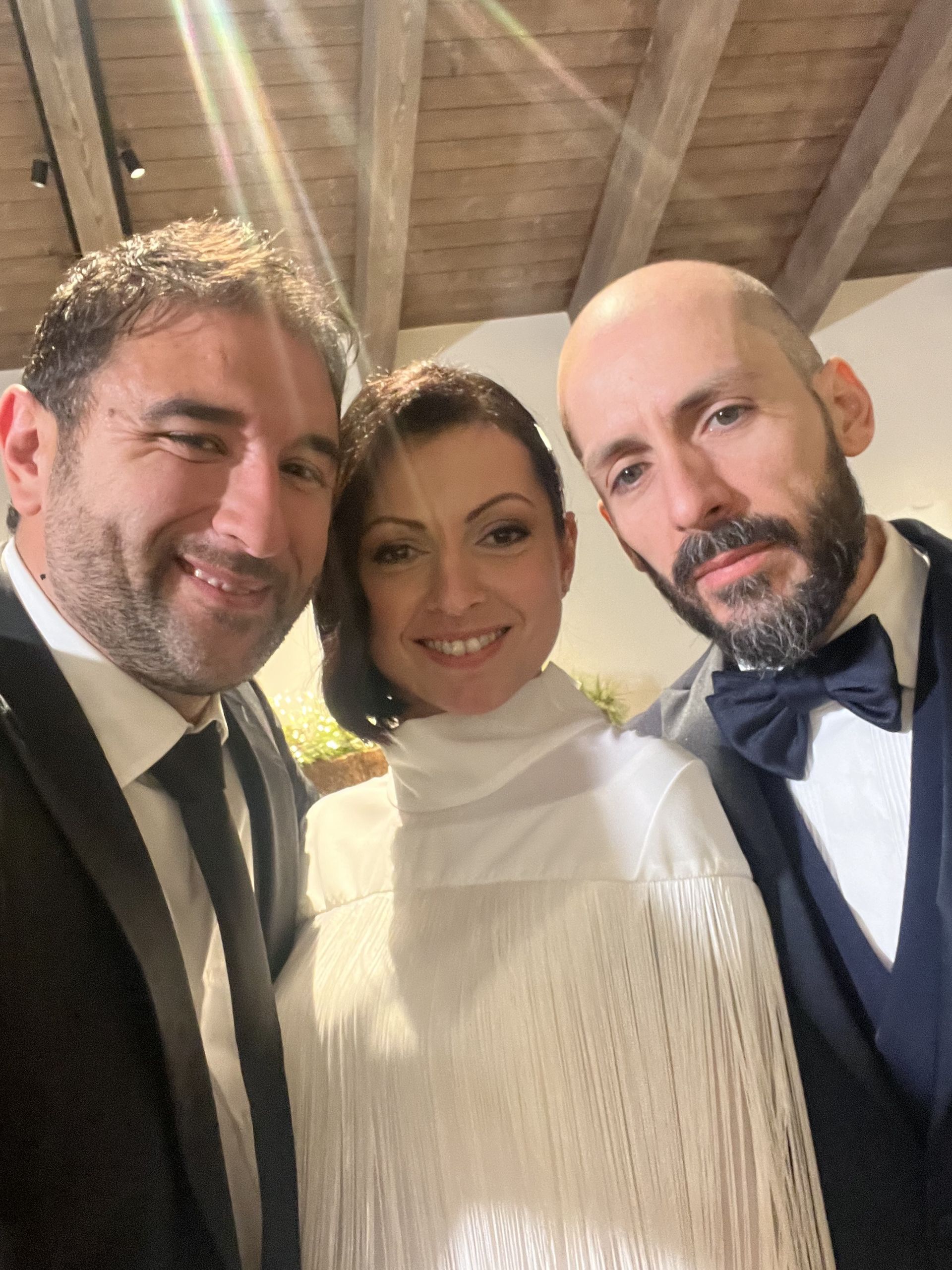 Tre persone sorridenti in un ambiente interno, sotto un soffitto di legno, vestite in abiti formali.