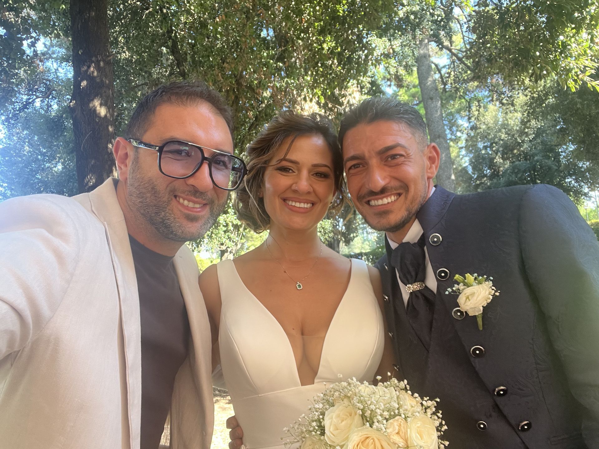 Tre persone sorridenti posano all'aperto, una delle quali tiene in mano un bouquet bianco, in un'ambientazione da matrimonio.