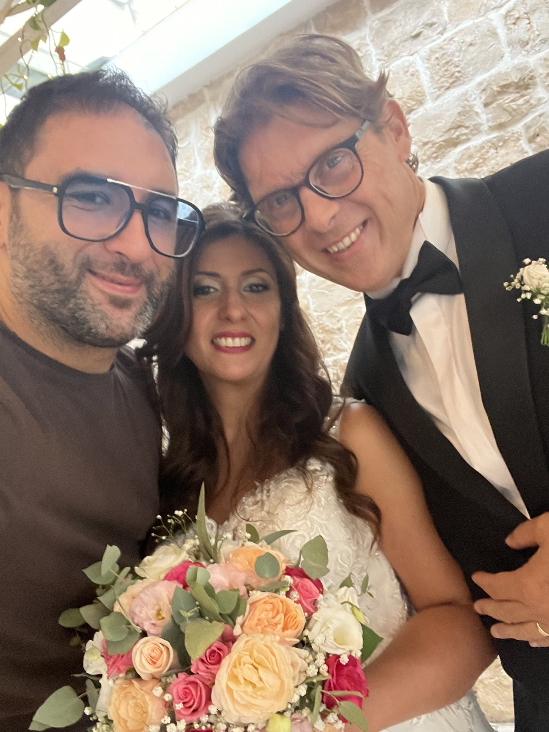 Tre persone sorridenti a un matrimonio, con la sposa che tiene in mano un bouquet dai colori pastello.