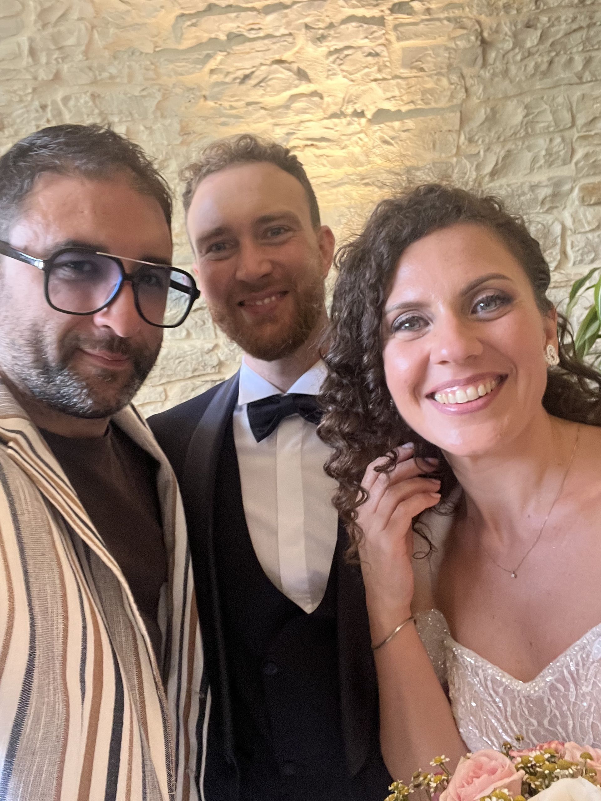 Tre persone sorridenti in abiti formali posano in un ambiente interno, davanti a un muro di pietra.