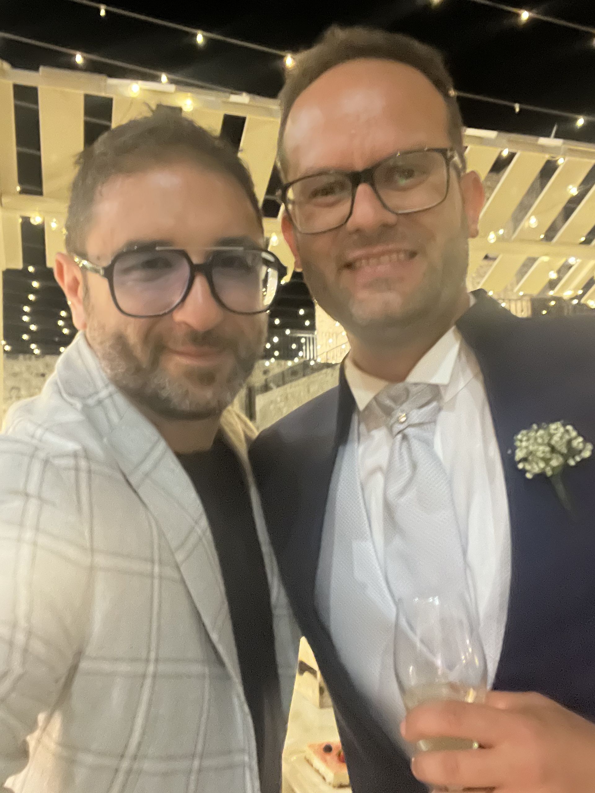 Due persone con gli occhiali sorridono a un evento serale formale, una delle due tiene in mano un drink.