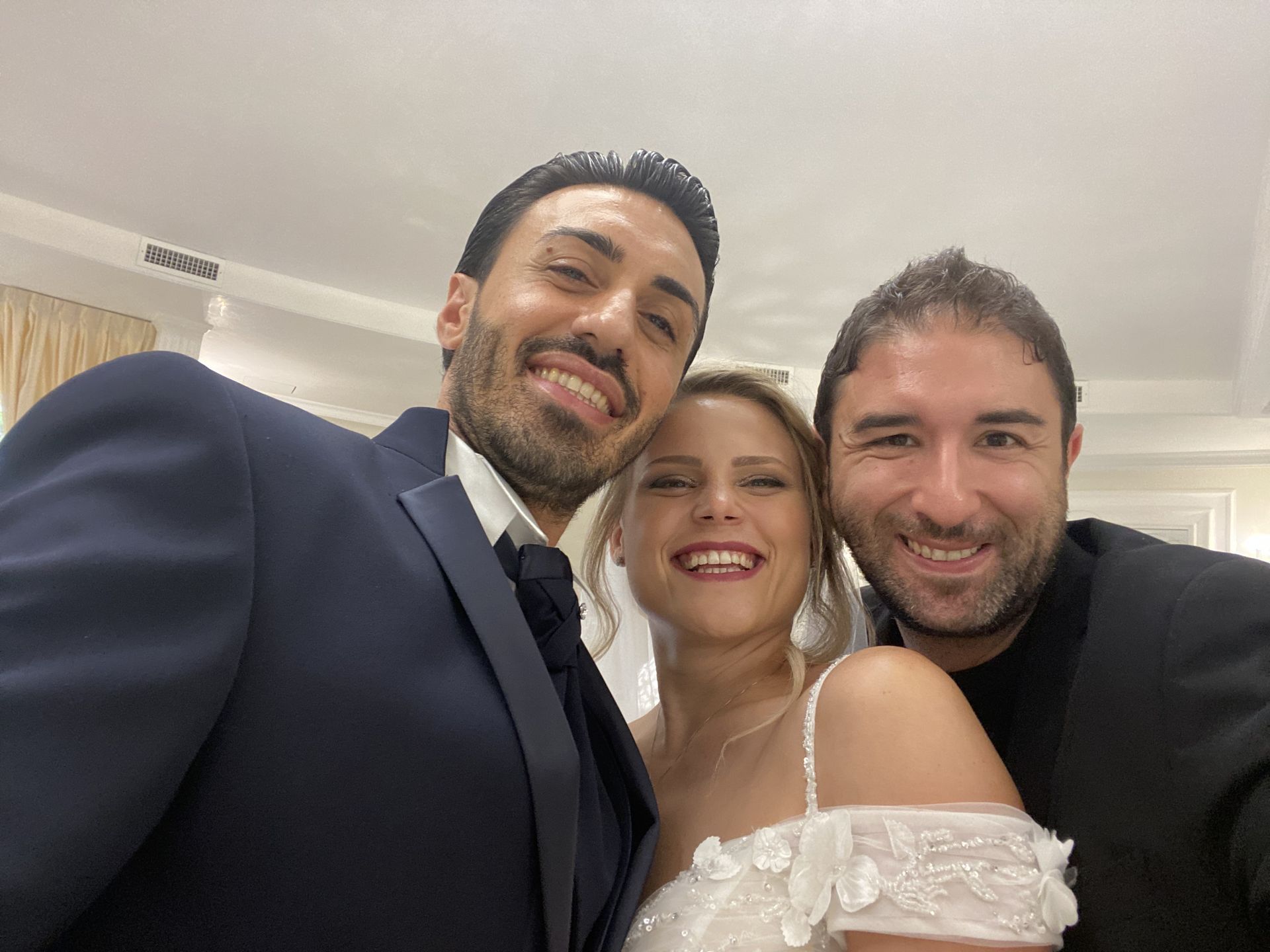 Tre persone sorridenti e vicine, in un ambiente interno, vestite in abiti formali, con la persona al centro che indossa un abito bianco.
