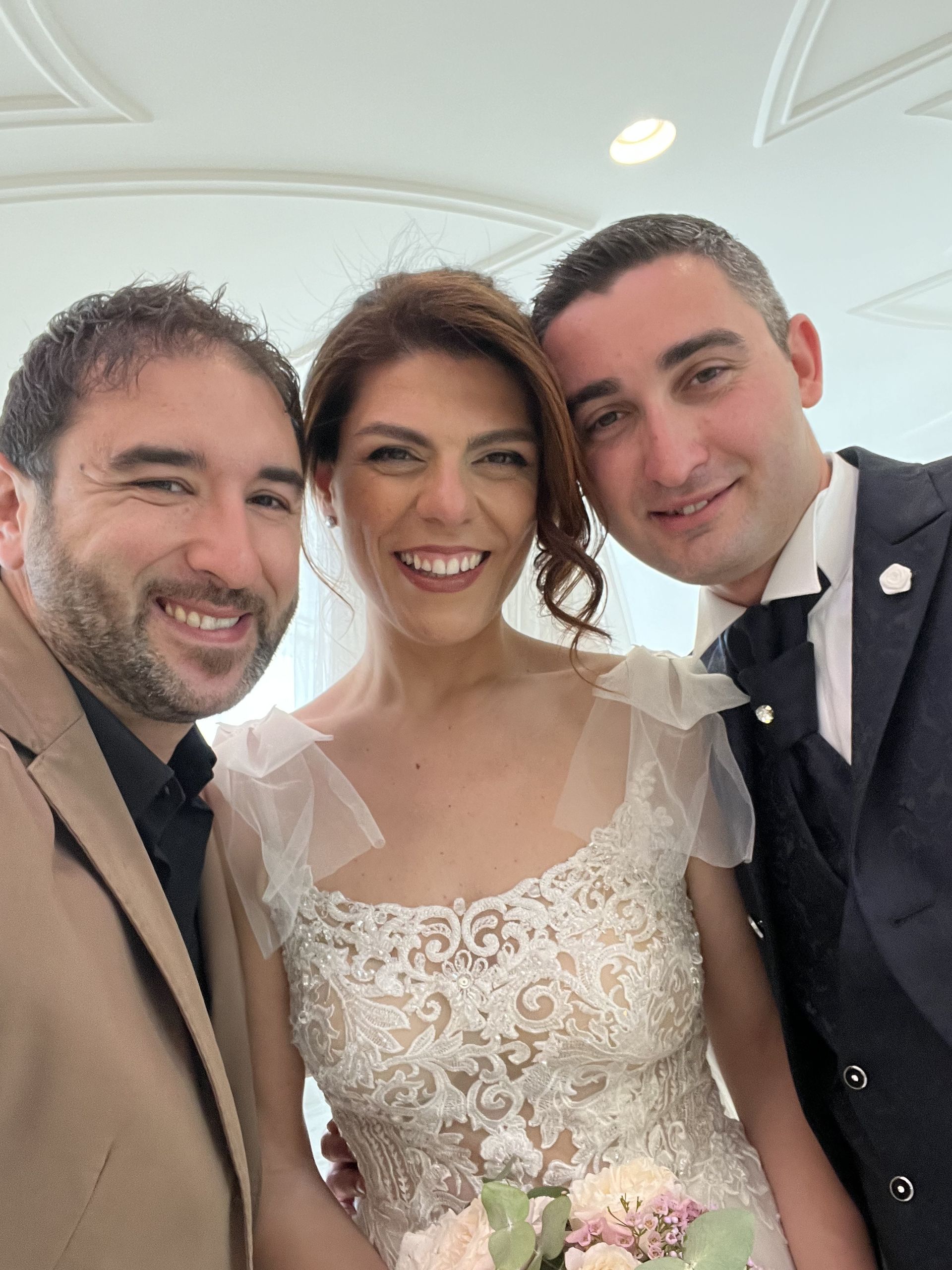 Tre persone sorridenti posano a un matrimonio, con la sposa in un abito di pizzo bianco che tiene in mano un bouquet.