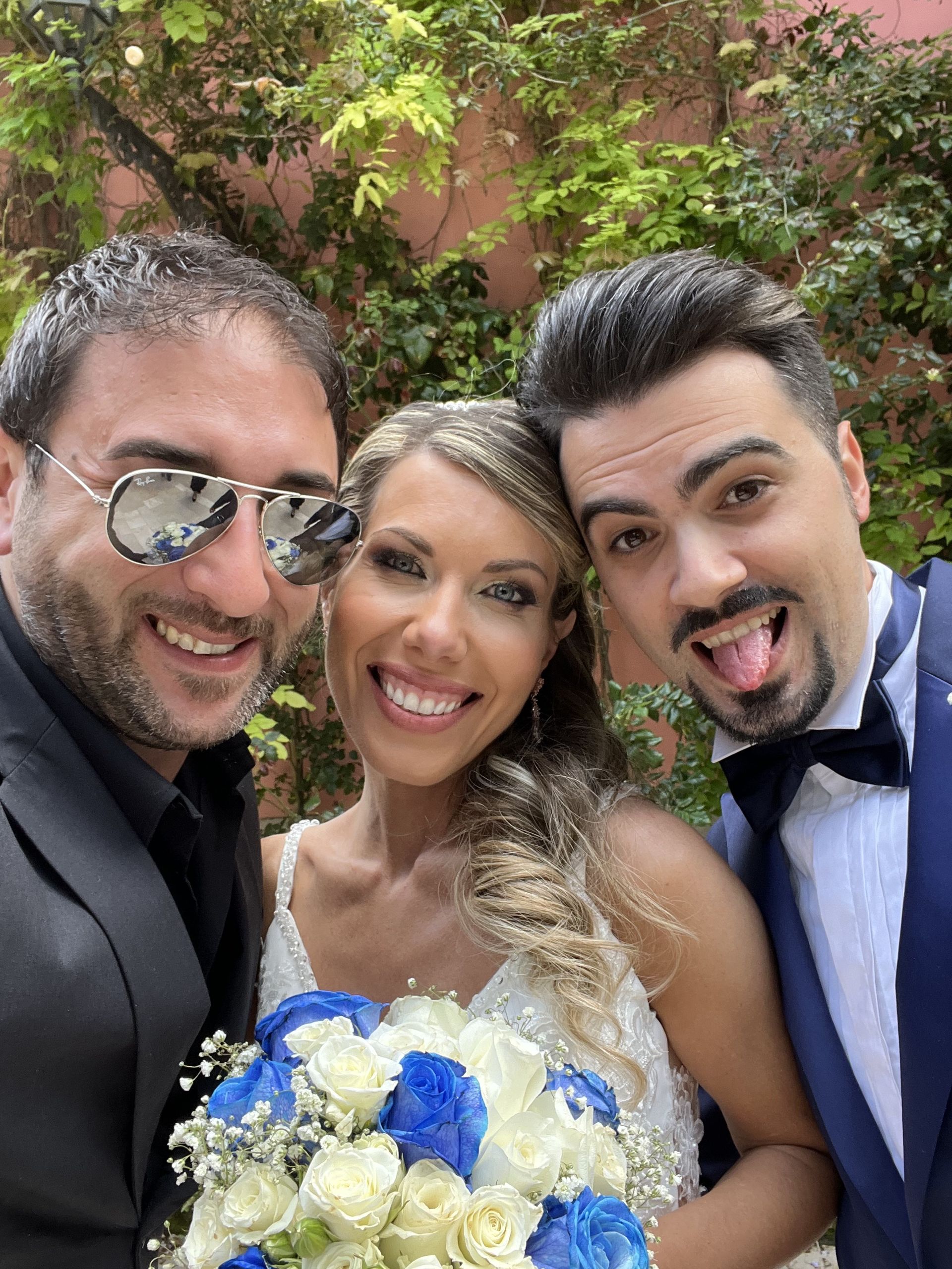 Tre persone sorridenti in abito elegante si scattano un selfie all'aperto, con un bouquet blu e bianco.