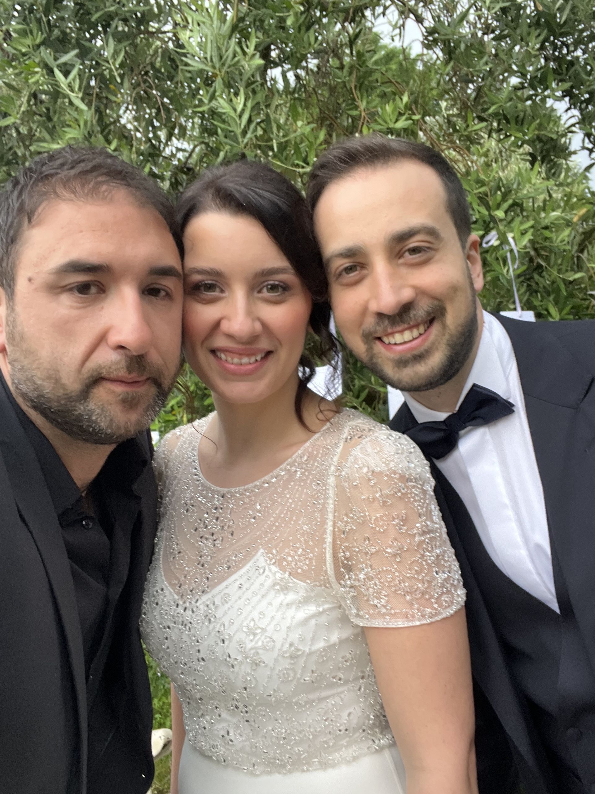 Tre persone sorridenti all'aperto, con la persona al centro che indossa un abito bianco e le altre in abiti eleganti scuri.